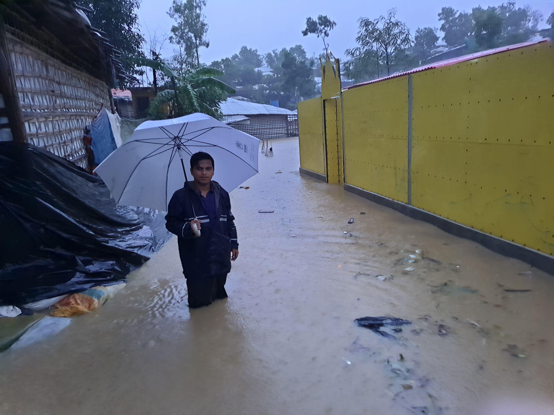 Person standing in knee-deep floodwater holding an umbrella near buildings and trees.