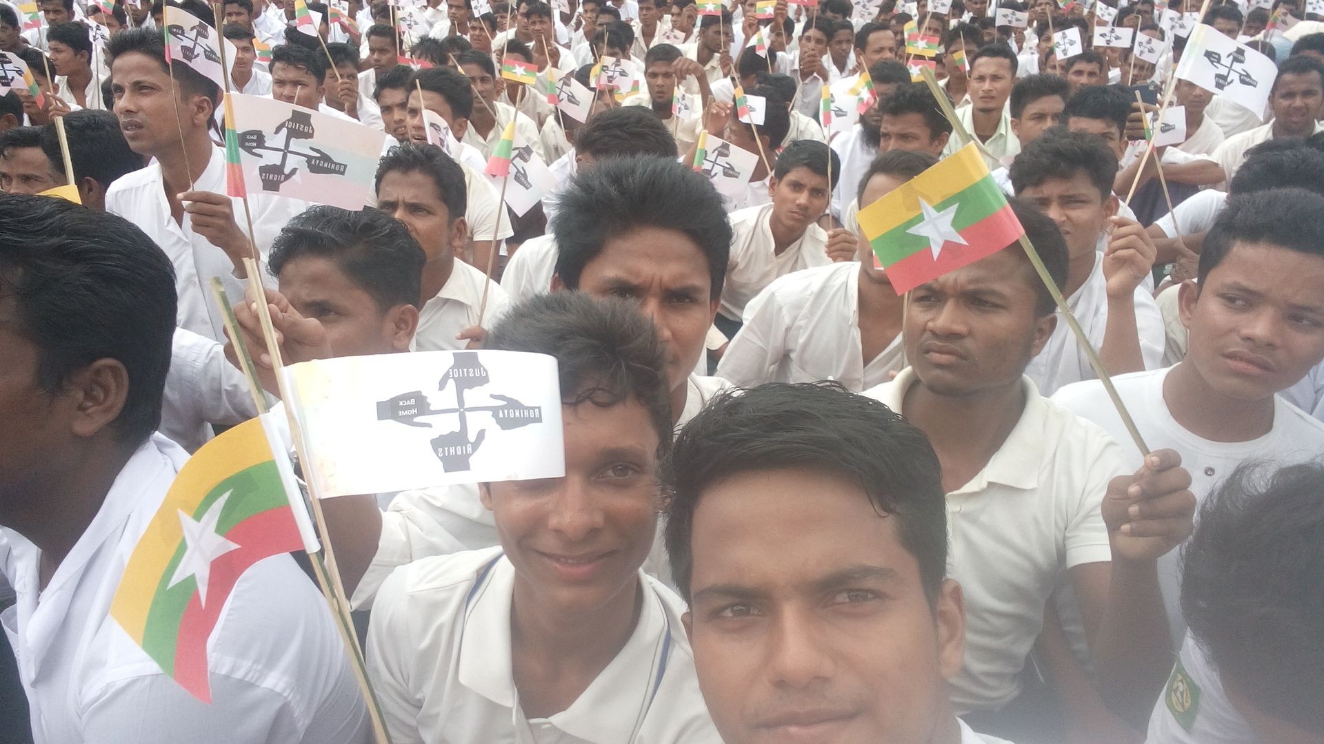 Group of men in white shirts holding flags with symbols and colors, gathered closely together.