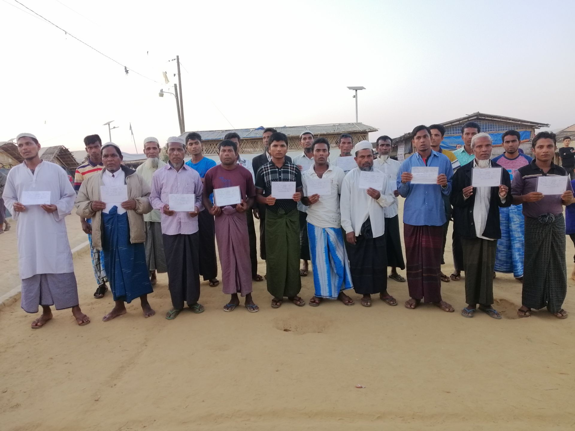 Group of people standing in a sandy area holding certificates with small buildings in the background.